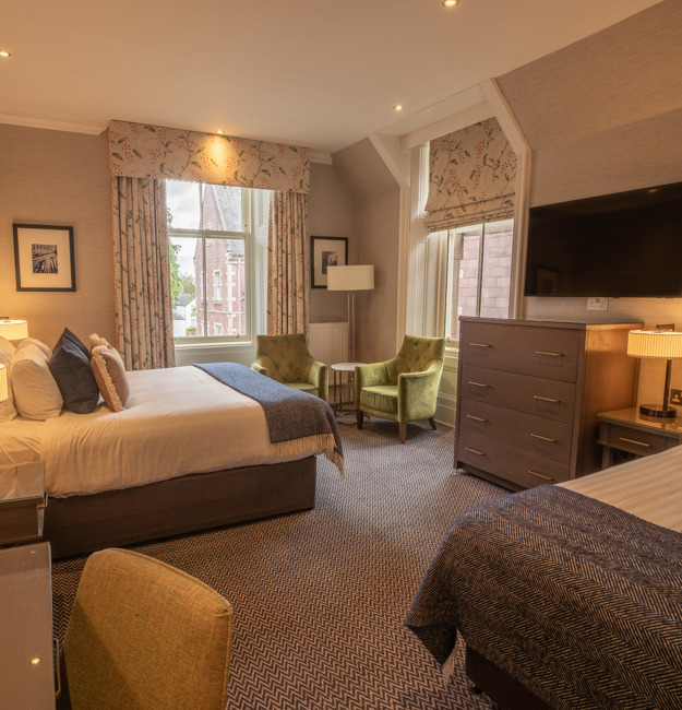 Room for 3 with a king-sized bed, a single bed on the opposite wall, two chairs by two windows, a dresser, a TV, and a vanity.