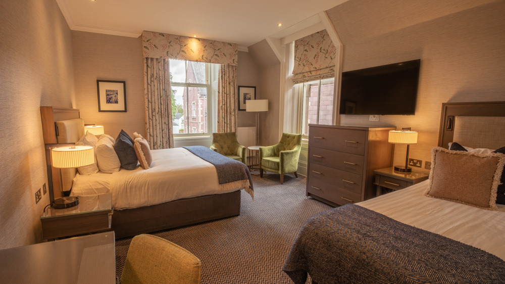 Room for 3 with a king-sized bed, a single bed on the opposite wall, two chairs by two windows, a dresser, a TV, and a vanity.