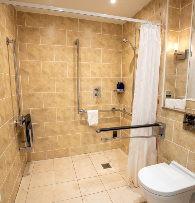 Accessible bathroom with roll-in shower with handles, toilet, sink, and towel rack.