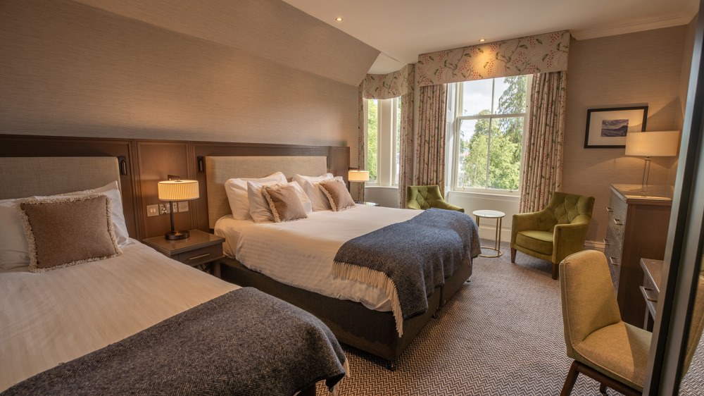 Room for 3 with a king-sized bed, a single bed, a vanity wit chair, dresser, and two chairs next to a window.