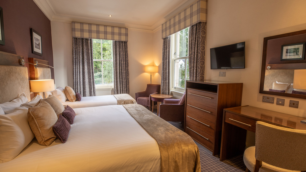 Room for 3 with a king-sized bed, a single bed, a vanity wit chair, dresser, TV on the wall, two chairs and two windows.